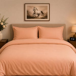 Può includere: Un set copripiumino color pesca a tinta unita con due federe rettangolari abbinate. La biancheria da letto è disposta su un letto con testiera dai toni neutri, affiancato da due comodini in legno, un vaso di fiori e una lampada.