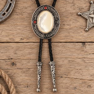 May include: A bolo tie featuring an oval silver-toned metal centerpiece with an inlaid iridescent white stone. The frame is decorated with small red and green accents. Two black braided leather cords hang down, finished with ornate silver-toned metal tips.