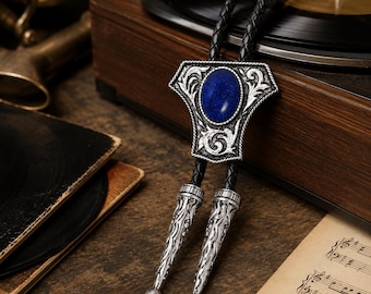 Puede incluir: Una corbata bolo con una piedra central ovalada de lapislázuli azul engastada en un marco de metal plateado adornado con intrincados motivos de volutas. La corbata incluye un cordón de cuero negro trenzado con dos puntas de metal plateado a juego.