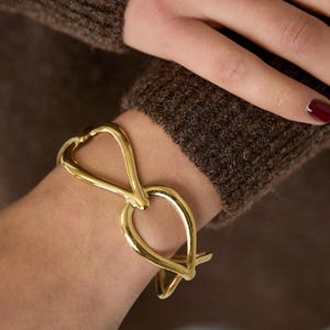 Peut inclure: Un bracelet manchette en métal doré présentant un design en boucle fluide inspiré de l'infini. Le bracelet possède une surface polie et réfléchissante ainsi qu'une structure lisse et arrondie qui entoure le poignet de manière symétrique.