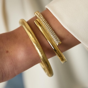 Può includere: Due bracciali color oro. Uno presenta una finitura opaca e testurizzata con un profilo arrotondato. Il secondo è un bracciale rigido color oro, liscio e lucido, con un elemento a barra rettangolare incastonato di piccole pietre chiare.
