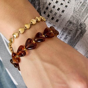 Pode incluir: Duas pulseiras e um anel são usados na mão. Uma pulseira apresenta pequenos elos dourados em forma de coração. A segunda pulseira consiste em contas maiores em forma de coração, de cor castanho escuro translúcido. O anel é uma banda dourada.
