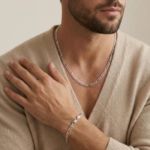 Może przedstawiać: Srebrny naszyjnik z łańcuszkiem i pasująca bransoletka. Biżuteria ma klasyczny design z ogniwami typu curb. Naszyjnik jest na szyi, a bransoletka na nadgarstku. Tło jest neutralne beżowe.