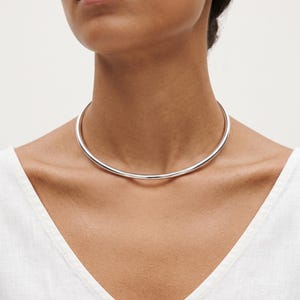 Peut inclure: Un collier ras-du-cou en métal argenté poli, doté d'une bande circulaire lisse, arrondie et continue qui repose fermement à la base du cou. La pièce présente une finition réfléchissante très brillante et un design simple et épuré.