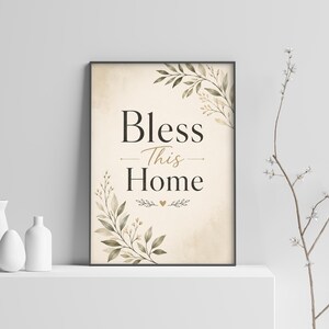 Op de afbeelding: Een rechthoekige wandkunstprint in een zwarte lijst met de tekst Bless This Home in een donkerbruin schreeflettertype. De achtergrond is zacht crèmekleurig met aquarelachtige groene en bruine botanische bladillustraties en een klein hartje.