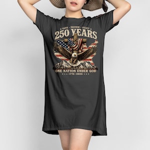 Puede incluir: Un vestido tipo camiseta negro con un estampado gráfico de un águila calva, una cruz y un mapa con la bandera estadounidense. El texto dice 250 YEARS, FAITH FREEDOM LEGACY, ONE NATION UNDER GOD y 1776-2026. Incluye siluetas de montañas en la base.