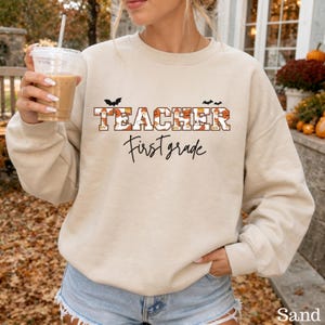 Puede incluir: Una sudadera beige de cuello redondo con la palabra TEACHER en letras de bloque rellenas con un patrón de calabazas y fantasmas, con pequeños murciélagos negros encima. Debajo, el texto First grade está escrito en cursiva negra.