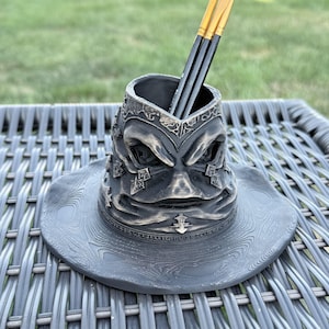 May include: A dark gray, weathered-looking desk organizer shaped like a pointed hat with a wide brim. The surface features intricate, raised textures and folds. Two thin paintbrushes with orange bristles and black handles are placed inside the top.