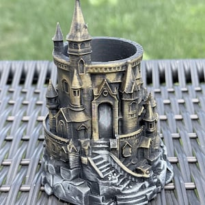May include: A cylindrical desk organizer shaped like a miniature castle with a black and metallic gold finish. The structure features multiple towers, a central arched doorway, and a staircase leading up to the entrance, all set on a rocky base.