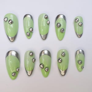 Puede incluir: Un juego de diez uñas postizas en forma de almendra con un acabado verde claro brillante. Cada uña presenta una punta plateada metálica y está decorada con pequeños tachones esféricos plateados en relieve sobre la superficie.