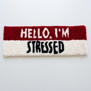 May include: A rectangular rug with a red top section and a white bottom section. The text "HELLO. I'M" is in white on the red section, and "STRESSED" is in black on the white section. The rug has a soft, textured appearance.
