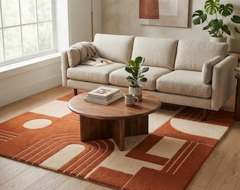 May include: A rectangular area rug featuring a geometric pattern in burnt orange and cream. The design includes concentric arches, circles, and rectangular blocks. The rug is placed on a light wood floor beneath a round wooden coffee table.
