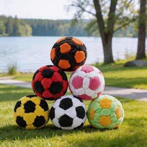 May include: A stack of colorful plush soccer balls in various colors, including black and orange, red and black, pink and white, yellow and black, white and black, and green and orange. The balls are on a grassy surface with a lake and trees in the background.