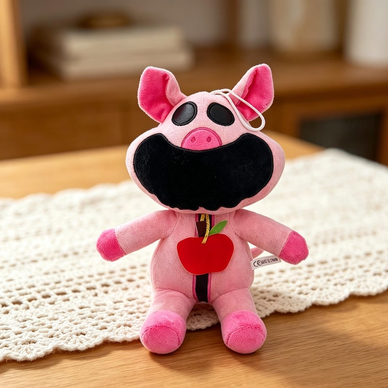 Puede incluir: Un peluche rosa con forma de cerdo con ojos negros y boca negra. El cerdo tiene orejas, brazos y pies rosas. Una manzana roja con una hoja verde est&aacute; unida al frente. Una cuerda blanca est&aacute; unida a la parte superior de la cabeza del cerdo.
