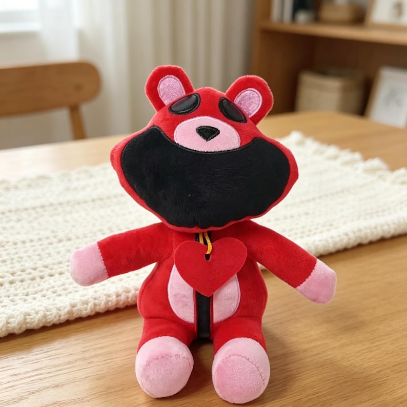Puede incluir: Un oso de peluche rojo con cara negra, orejas y pies rosas, y un colgante de coraz&oacute;n rojo. El oso tiene una cuerda amarilla alrededor del cuello y est&aacute; sobre una mesa de madera. Es un peluche.