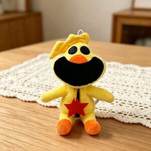 Puede incluir: Un peluche amarillo con boca negra, dos ojos negros y pies naranjas. El juguete tiene una estrella roja en el pecho y un lazo amarillo en la cabeza. El juguete est&aacute; sobre una superficie blanca y de encaje.