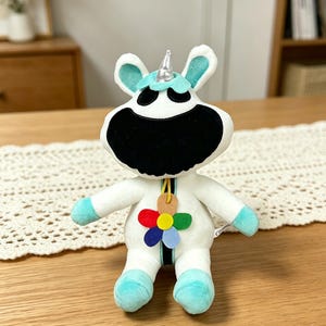 Puede incluir: Un peluche de unicornio con cuerpo blanco, cara negra y detalles turquesa en las orejas y los pies. Tiene un cuerno plateado y un colgante de flores de colores. El juguete est&aacute; sobre una mesa de madera.
