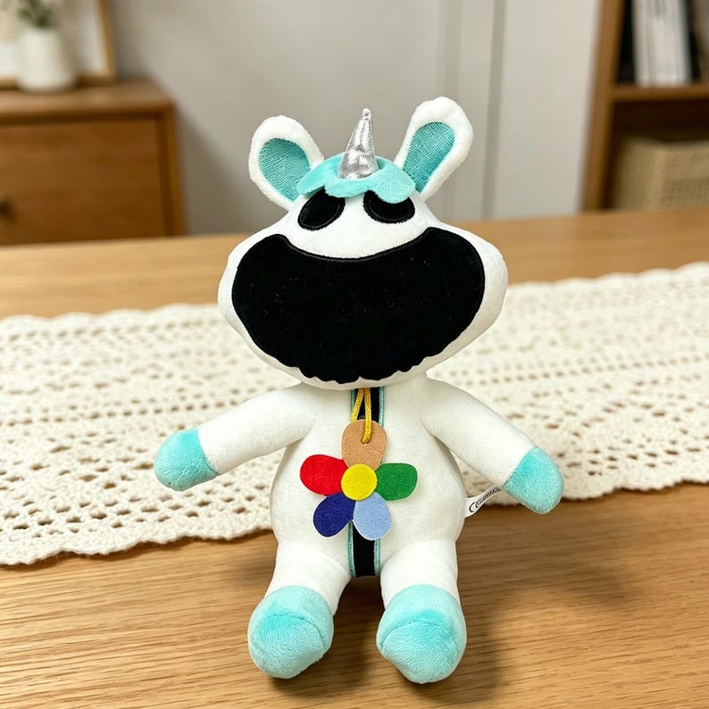 Puede incluir: Un peluche de unicornio con cuerpo blanco, cara negra y detalles turquesa en las orejas y los pies. Tiene un cuerno plateado y un colgante de flores de colores. El juguete est&aacute; sobre una mesa de madera.