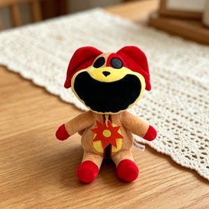 Puede incluir: Un peluche con forma de personaje de dibujos animados con cabeza y orejas rojas, cara amarilla y boca grande negra. El juguete tiene cuerpo color canela, manos y pies rojos, y un emblema en forma de estrella en el pecho. El juguete est&aacute; sobre una superficie de madera.