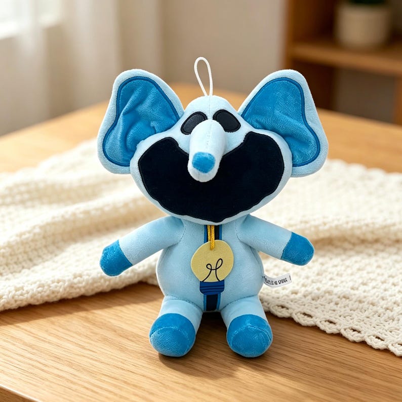 Puede incluir: Un peluche de elefante azul claro con grandes orejas azules y una cara negra. El elefante tiene un colgante de bombilla y una cuerda blanca para colgarlo. El juguete est&aacute; sobre una superficie de madera.