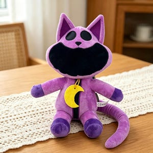 Puede incluir: Un peluche de gato morado con boca y ojos negros, y un colgante de media luna amarilla. El gato tiene una cola larga y detalles morados en las patas y orejas. El juguete est&aacute; sobre una mesa.