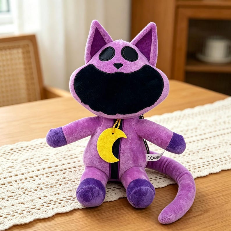 Puede incluir: Un peluche de gato morado con boca y ojos negros, y un colgante de media luna amarilla. El gato tiene una cola larga y detalles morados en las patas y orejas. El juguete est&aacute; sobre una mesa.