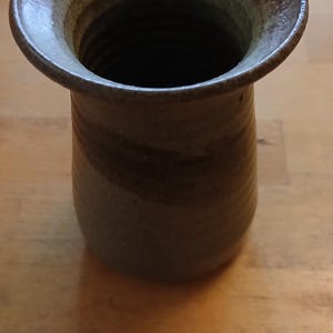 May include: A cylindrical stoneware vase with a wide, flared rim. The exterior features a mottled, earthy colour palette with blended tones of brown, grey, and tan. The surface has a visible, textured finish with subtle horizontal ridges along the body.