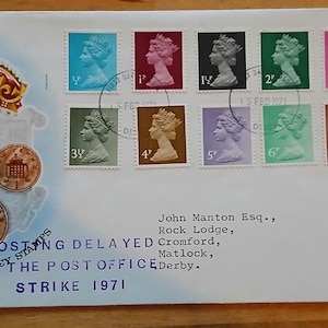 May include: A white first day cover envelope featuring a collection of twelve colourful postage stamps with Queen Elizabeth II profiles. The envelope displays a crown illustration, four coin images, and text reading POSTING DELAYED BY THE POST OFFICE STRIKE 1971, addressed to John Manton Esq. in Cromford, Matlock, Derby.