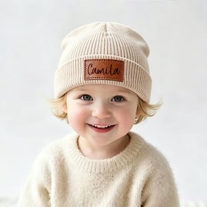 May include: A cream-colored ribbed knit beanie featuring a rectangular brown faux leather patch on the front. The patch is embossed with the name Camila in a dark brown script font. The beanie has a folded cuff and a textured knit construction.