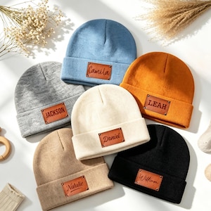 May include: Six knit beanies in blue, gray, cream, orange, tan, and black. Each beanie features a rectangular brown leather patch riveted to the cuff, engraved with the names Camila, Jackson, Daniel, Leah, Natalie, and William respectively.