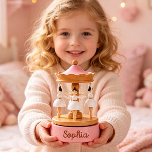 May include: A wooden musical carousel toy featuring three miniature ballerina figures in white dresses. The carousel has a pink roof and a pink base with the name Sophia engraved in a cursive script on the front of the circular wooden base.