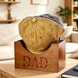 May include: A wooden box with the word "DAD" engraved on the front, holding several baseball caps. The caps are in shades of tan, blue, and gray. The box also has names engraved below the word "DAD".