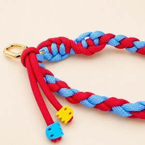 Può includere: Un cinturino in corda intrecciata con fili di nylon rosso e azzurro alternati. Il cinturino è fissato a un anello a D in metallo dorato e termina con due estremità di corda rossa, ciascuna rifinita con un piccolo ciondolo a forma di fantasma.