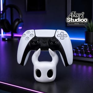 May include: A white and black PlayStation 5 controller on a white stand. The stand has a distinctive design with curved horns and eye-shaped openings. The Akari Studioo logo is visible in the background, with a keyboard and headphones.
