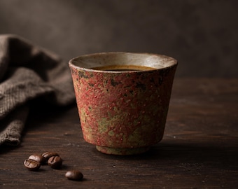 May include: A rustic ceramic coffee cup with a textured red and green exterior, filled with dark coffee. The cup sits on a dark wooden surface with coffee beans and a folded cloth in the background.