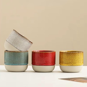 Op de afbeelding: Vier keramische bekers in een stapel, elk met een unieke kleur en textuur. De bekers hebben een tweekleurig ontwerp, met een lichtbeige basis en een bovenste gedeelte in teal, rood, geel en wit. De bekers zijn cilindrisch met afgeronde bodems.