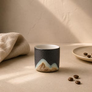 Può includere: Una piccola tazza cilindrica in ceramica con esterno nero strutturato. Il fondo della tazza presenta un disegno a forma di montagna nei toni del marrone e dell'azzurro. L'interno è bianco. Chicchi di caffè sparsi nelle vicinanze.