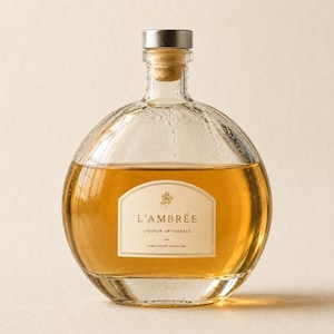 Peut inclure: Une bouteille en verre transparente, de forme arrondie, remplie d'un liquide ambré. Elle est fermée par un bouchon en liège et un capuchon métallique. L'étiquette blanche porte l'inscription "L'AMBRÉE, Liqueur Artisanale, Fabrication Française".