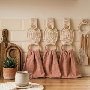 May include: Three cream-colored, crocheted kitchen towel holders with wooden rings and buttons, each holding a pink towel with white polka dots. A mug, cutting boards, and a plant in a terracotta pot are also visible.