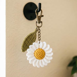 Op de afbeelding: Een gehaakte sleutelhanger met een witte madeliefbloem met een geel hart en een enkel olijfgroen blaadje. De bloembedel is bevestigd aan een bronskleurige metalen karabijnhaak en een korte ketting, hangend aan een zwarte wandhaak.
