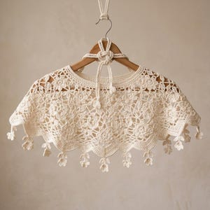 Può includere: Una mantellina all'uncinetto color bianco sporco caratterizzata da un intricato motivo in pizzo floreale con orlo smerlato. Il capo presenta piccoli motivi floreali pendenti lungo il bordo inferiore e una chiusura centrale a laccio sul collo.