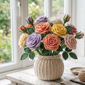 Op de afbeelding: Een handgemaakt boeket gehaakte rozen in diverse kleuren, waaronder perzik, geel, lavendel en roze, gerangschikt in een crèmekleurige, geribbelde gehaakte vaas. De bladeren zijn groen en het arrangement staat op een witte houten tafel bij een raam.