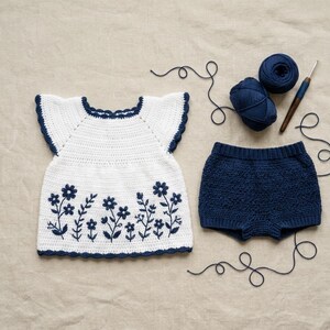 May include: A white crocheted top with short flutter sleeves and a dark blue floral pattern along the hem, paired with matching dark blue crocheted shorts. Two balls of dark blue yarn and a wooden crochet hook are positioned next to the garments.