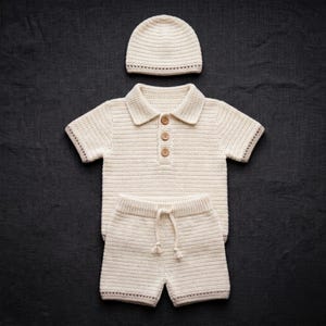 May include: A cream-colored crochet set consisting of a short-sleeved polo shirt with three brown buttons, a pair of shorts with a drawstring waist, and a matching beanie hat. The items feature a consistent textured knit pattern throughout.