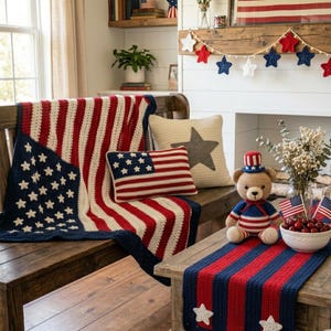 May include: A crochet American flag blanket, a rectangular flag pillow, a square pillow with a grey star, and a crochet teddy bear wearing a patriotic hat. A red, white, and blue striped table runner with white stars sits on a wooden table.