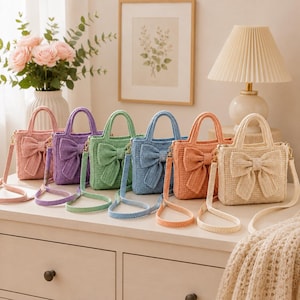 May include: A row of six small, woven tote bags in pink, purple, light green, blue, orange, and cream. Each bag features a large, matching woven bow on the front, two top handles, and a long, thin shoulder strap attached with gold-tone hardware.