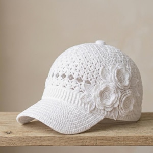 Peut inclure: Une casquette de baseball blanche en fil crocheté, dotée d'une visière structurée et d'une calotte au motif ajouré complexe. Le côté de la casquette est orné d'applications florales et de feuilles en crochet en trois dimensions.