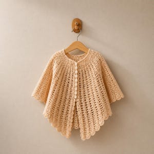 May include: A light peach-colored crochet capelet featuring a vertical ribbed pattern, a round neckline, and a front button closure with small matching buttons. The garment has a scalloped hemline and is displayed on a wooden hanger.