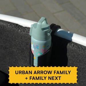 May include: A light blue water bottle with a floral pattern and a handle lid, secured in a black cup holder attached to a white bar. A yellow rectangular label with a black dashed border displays the text URBAN ARROW FAMILY + FAMILY NEXT.