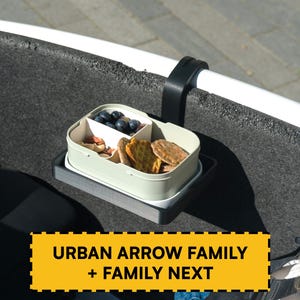 Può includere: Un contenitore per snack rettangolare verde chiaro con divisori interni contenente mirtilli e cracker, fissato a una staffa nera. Il testo giallo sovrapposto recita URBAN ARROW FAMILY + FAMILY NEXT in lettere maiuscole nere.